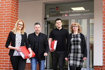 Vier Personen mit Aktenordnern in der Hand stehen vor einem Gebäude mit der Aufschrift 'Herzlich Willkommen'.