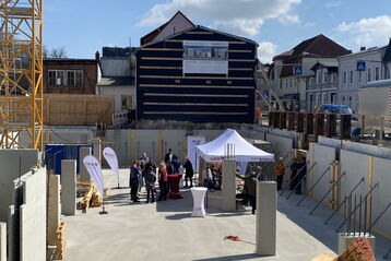 Baustelle mit mehreren Personen unter einem weißen Pavillon, umgeben von Bauwänden und einem teilweise verkleideten Gebäude im Hintergrund.