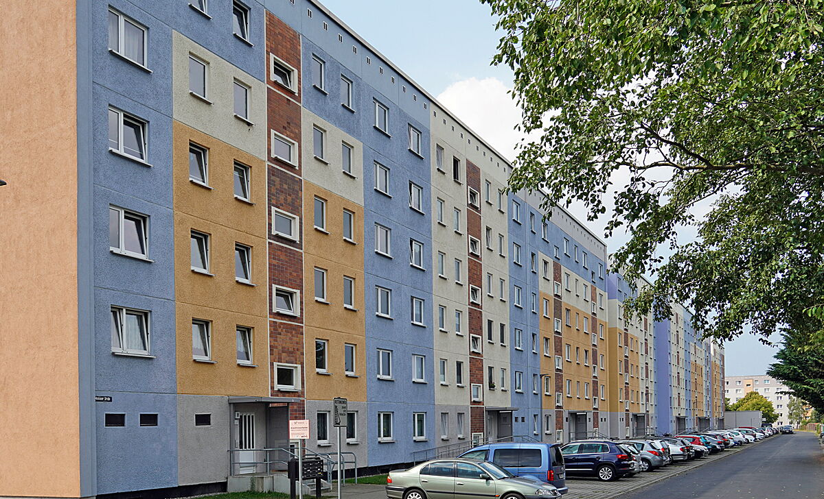 Langes Wohnhaus mit mehreren Stockwerken, farblich in Blau, Gelb und Weiß gestaltet, mit vielen Fenstern und geparkten Autos entlang der Straße, rechts Bäume und blauer Himmel.