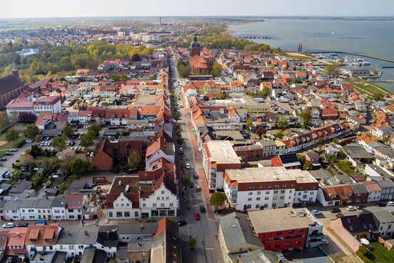 Luftaufnahme des Stadtzentrums von Ribnitz mit einer Hauptstraße, umgeben von dicht bebauten Häusern und im Hintergrund der Bodden mit Hafenanlagen.