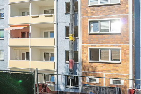 Mehrstöckiges Wohngebäude mit Bauabsperrung und einem langen, vertikal angebrachten Bauabfallrohr, das in einen Container mündet.