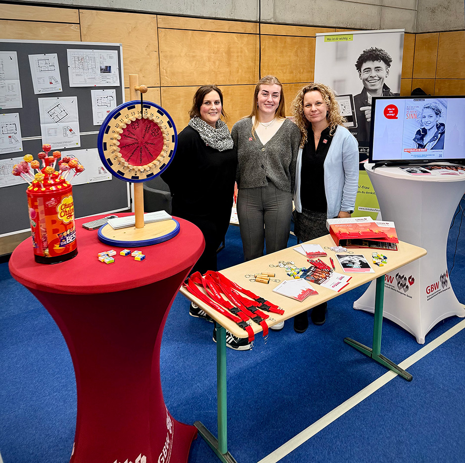 Drei Personen stehen hinter einem Tisch mit Werbematerialien und einem roten Stehtisch mit einem Glücksrad und einem Behälter mit Werbegeschenken in einem Raum mit blauer Bodenmatte und Holzpaneelen an der Wand.